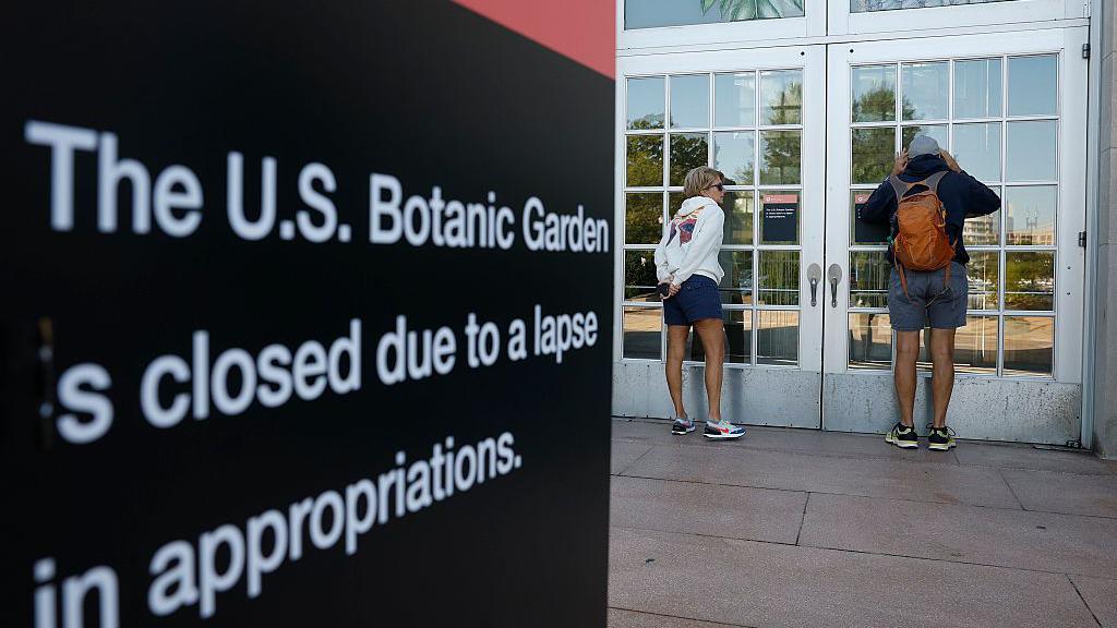 Two visitors in shorts and sweatshirts look through the door windows of the closed US Botanical Gardens during the federal government shut down. In the foreground is a black sign with white lettering reading: 'The US Botanic Garden is closed due to a lapse in appropriations'. 