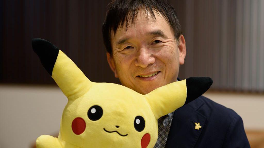 A man wearing a navy suit sits in an office, smiling. He's peeking over the top of a large, yellow plush version of Pokémon Pikachu.