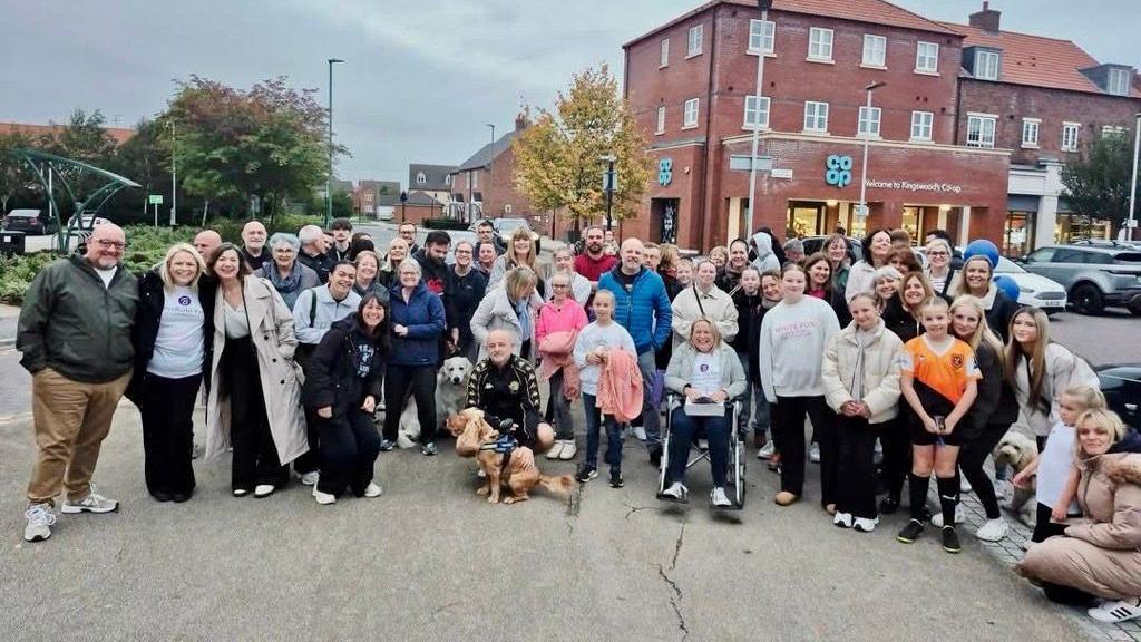 A large group of people are stood looking directly at the camera. There is a mixture of males, females, children and pets within the group. Some of them hug, lean on each other and wrap their arms around each other. They are stood in a car park, in front of a red bricked shop with many windows reading: 'Co Op' in blue and 'Welcome to Kingswood's Co op' in smaller white text.
