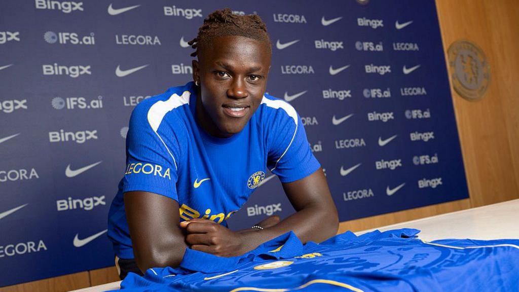 Quenda resting his arms on a table with a Chelsea shirt on and in front of him after signing for the club