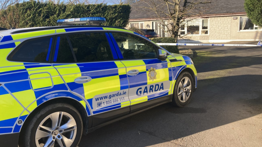 A Garda car in blue and yellow in front of a Garda tape and a bungalow