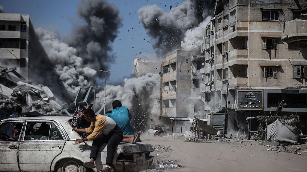 People in a car cower as a blast in the background causes a building to collapse in a huge plume of smoke and debris. Two people are also sitting on the back bonnet of the car.