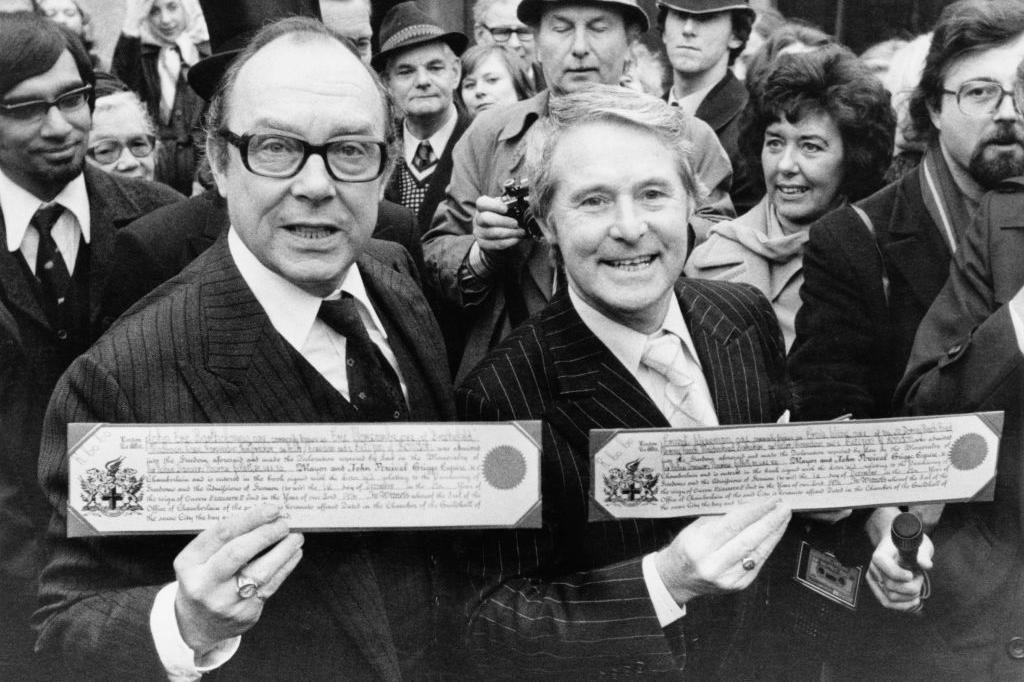 Black and white photo of Eric Morecambe and Ernie Wise holding their Freedom documents. They are both wearing suits and are surrounded by a crowd of people