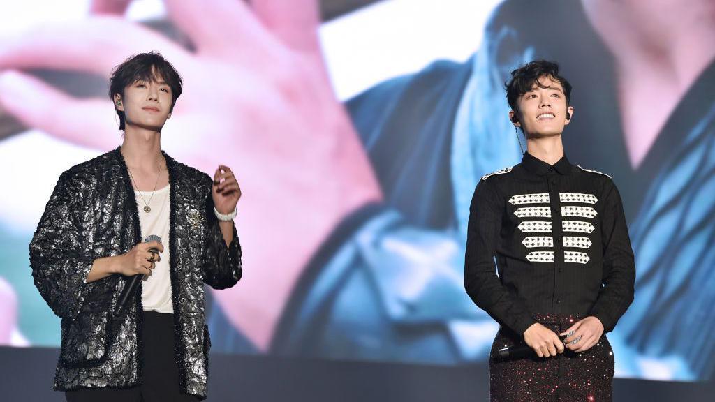 Actors Wang Yibo (L) and Sean Xiao Zhan (R) smiling on stage in a photo from November 1, 2019 in Nanjing. Wang is wearing a sequined jacket, white shirt and black pants, while Xiao is wearing a black shirt and sequined pants. Both are holding mics.