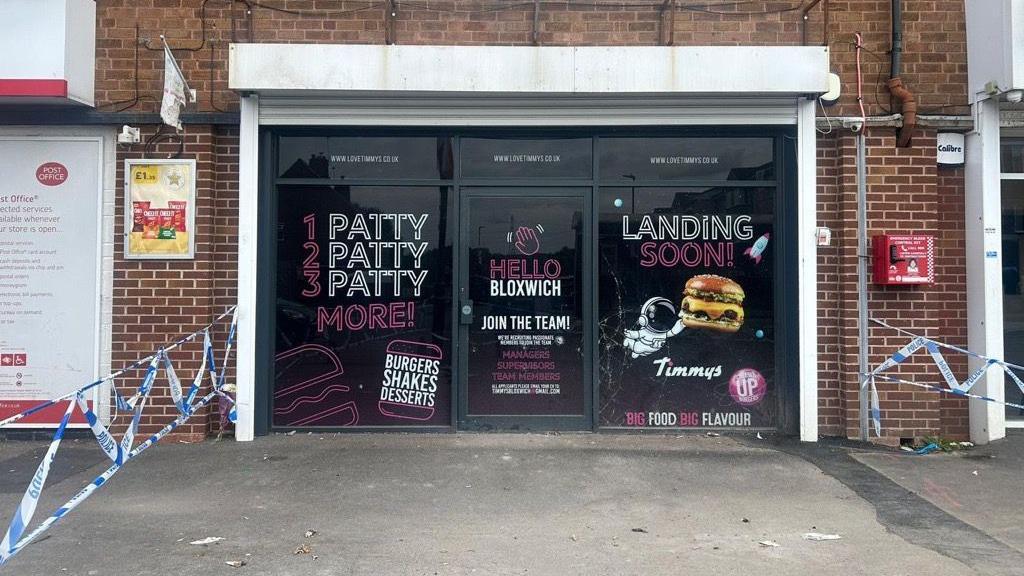 The front of the takeaway can be seen across the wide pavement outside. There are promotional posters stuck to the windows that tell people the burgers it will be producing are "landing soon". They also say "Hello Bloxwich". In the foreground police tape can be seen tied to a lamp-post.

