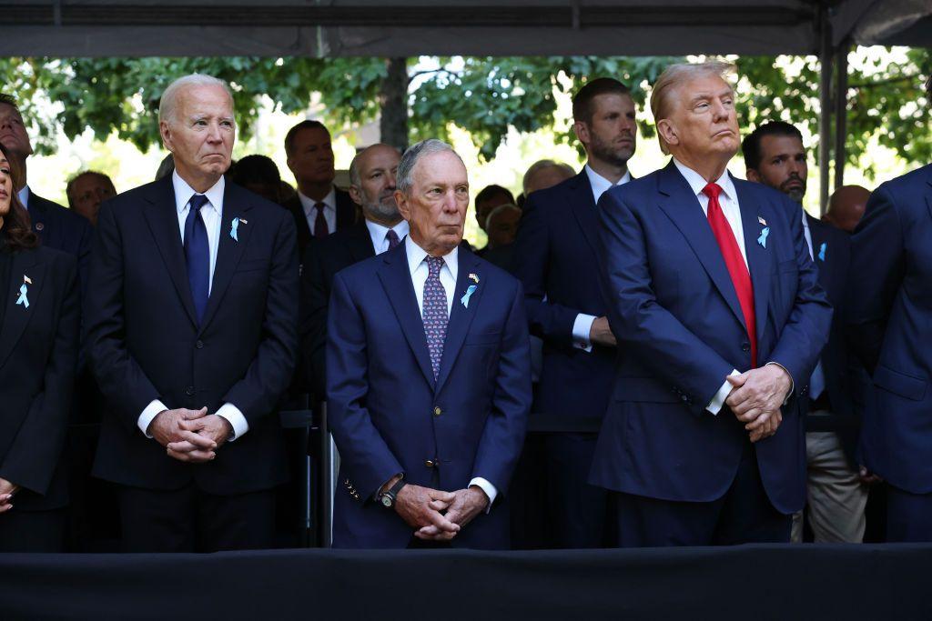 Joe Biden, Michael Bloomberg and Donald Trump stand in a line on the front row of a memorial service to remember the victims of the 9/11 attacks. 

There are several people stood behind them, all three have their hands clasped together in front of them. 