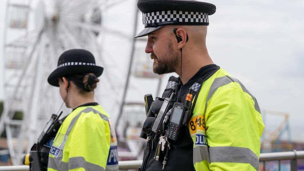 Two police officers wearing high visibility jackets and officer uniform, standing beside a fairground ride