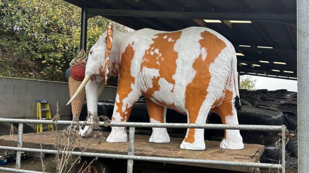 An orange elephant statue in a barn while it is midway through being repainted. It has orange and white stripes due to the work to remove the old paint.