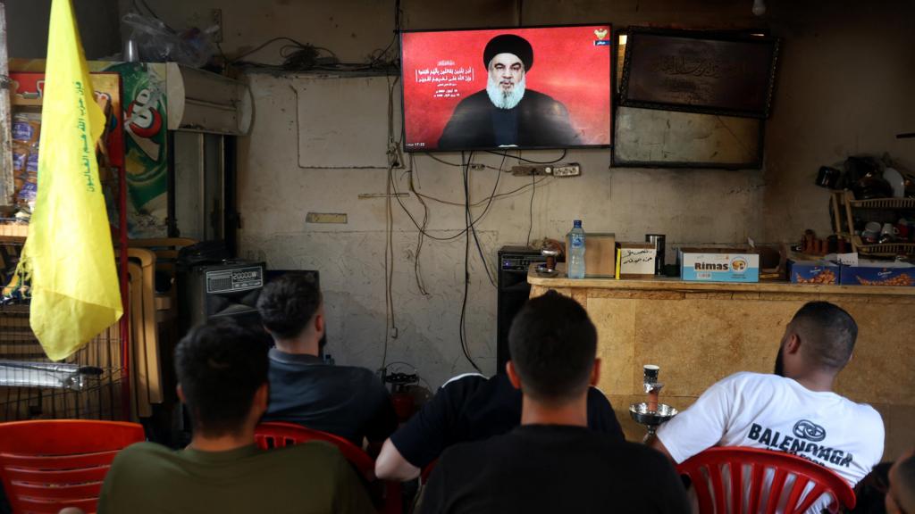 People in a cafe sit on red plastic chairs watching the Hezbollah leader give a speech on a wall-mounted TV