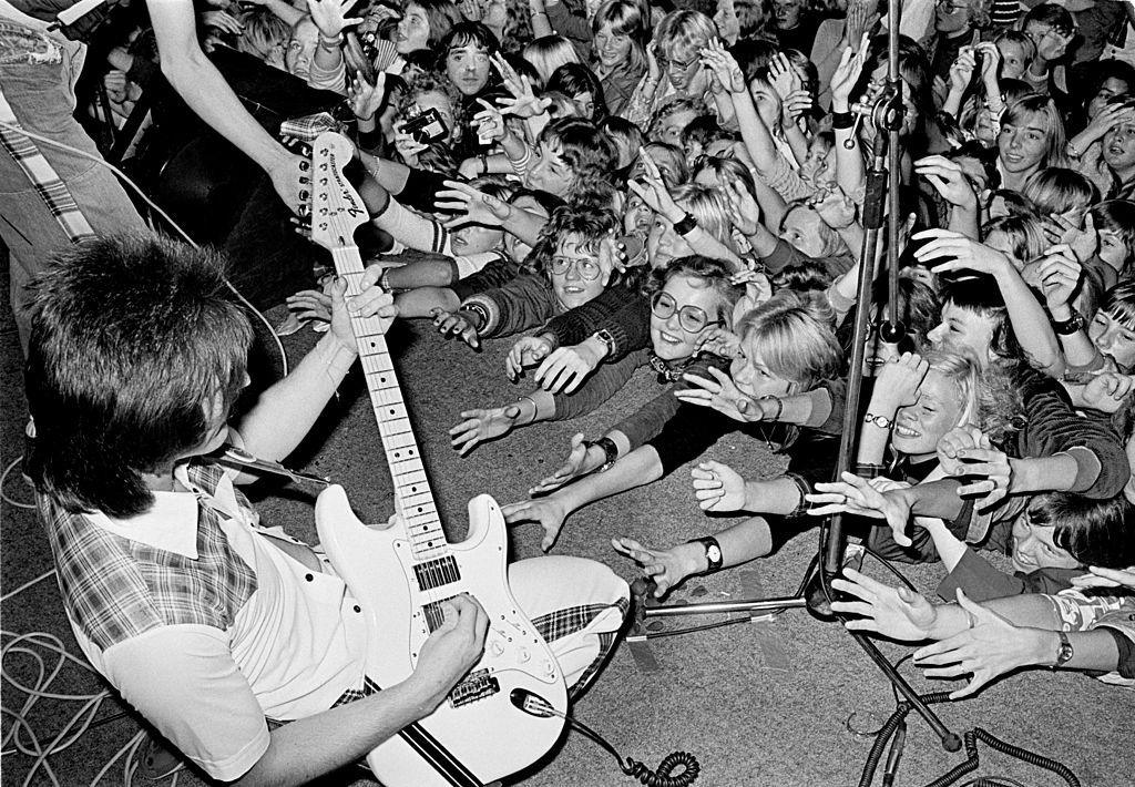 One of the band members plays guitar on stage, wearing a white suit. The shirt has tartan shoulders and the trousers have tartan stitching. Each person in the crowd has their arms outstretched and clawing at the boy on stage. 