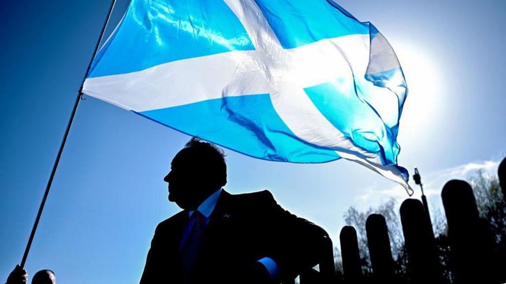 Salmond is standing in silhouette against a bright blue sky and weak winter sun, with a Saltire flying over his head. He is looking out to the left.