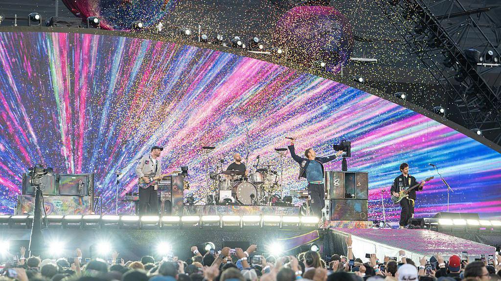 A live concert with four band members performing on stage under bright multicoloured lights and falling confetti.