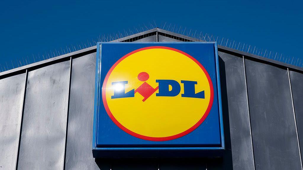 A large grey metal section of a building with a square blue sign. There is a red and yellow circle with the Lidl logo on the inside of it. The sky above the building is blue 