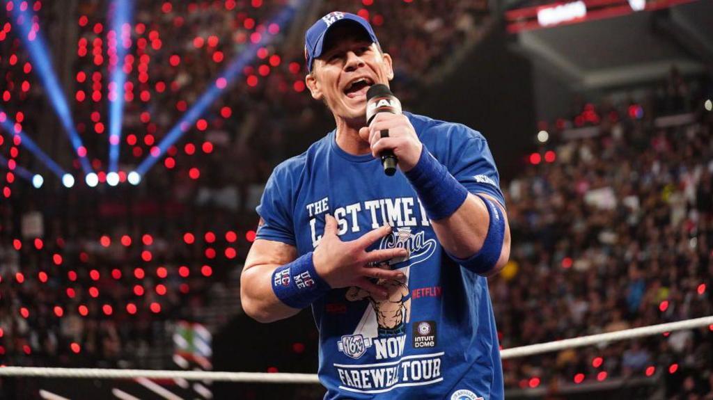 John Cena dressed in a blue t-shirt and cap stands in a ring talking into a microphone