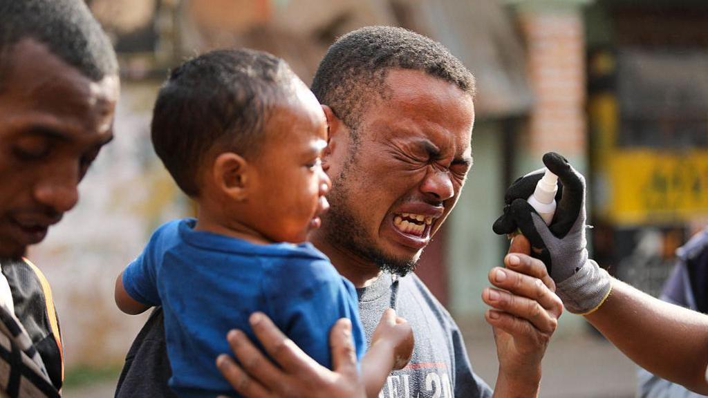 A man holding a child is grimacing as someone is spraying a liquid in his face after he was affected by tear gas.