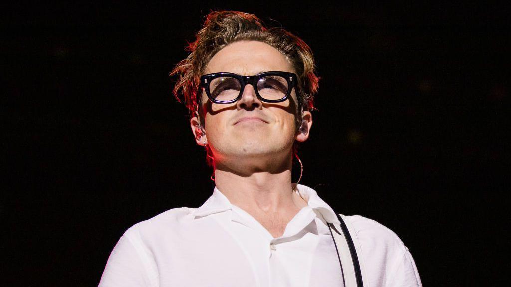 Head and shoulders shot of Tom Fletcher from McFly in a white top and thick black glasses