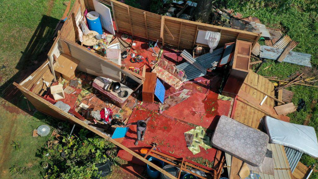 An aerial view of a wooden home, which is missing a roof. The wall on the left-hand side has fallen over, and inside the property various items are scattered across the floor. A woman can be seen close to what was the door, her arms held out wide as she assesses the damage.