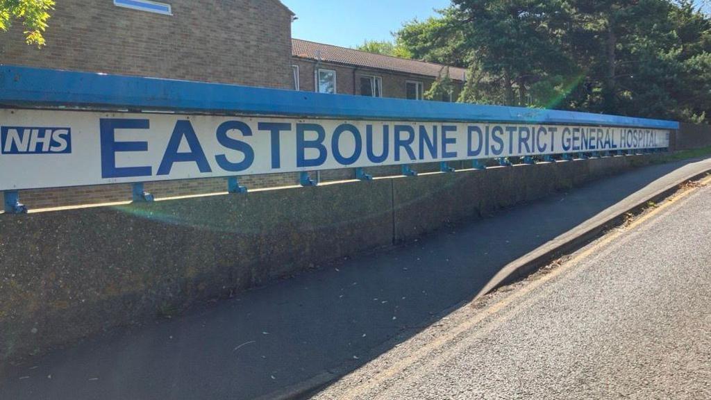 A blue sign on a white background reading "Eastbourne District General Hospital" with the NHS logo