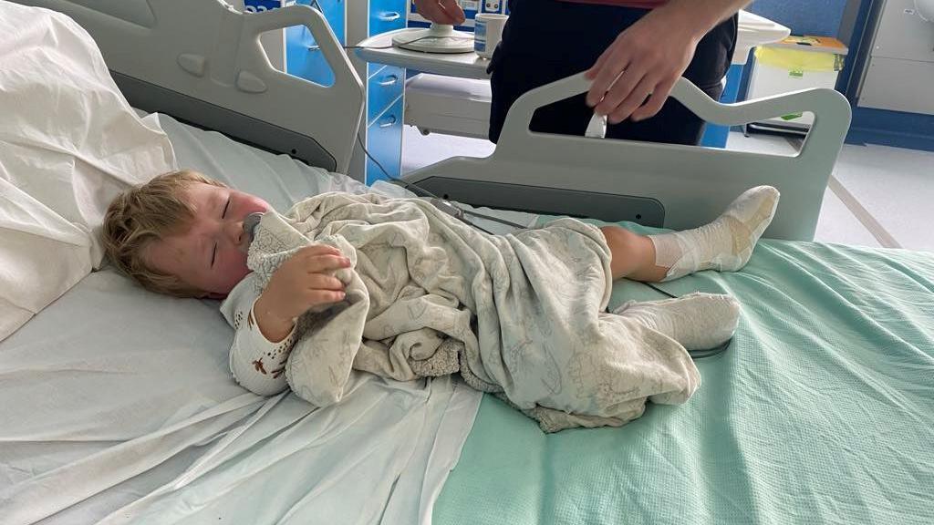 Edison is lying in a hospital bed, on top of green and white sheets. He has he eyes closed and is holding a blanket. His feet are wrapped in bandages.