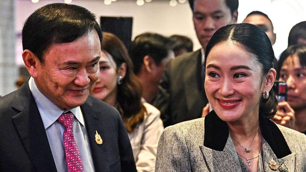 Thaksin and Paetongtarn smiling as they stand beside each other, Thaksin wearing a suit and Paetongtarn a grey blazer.