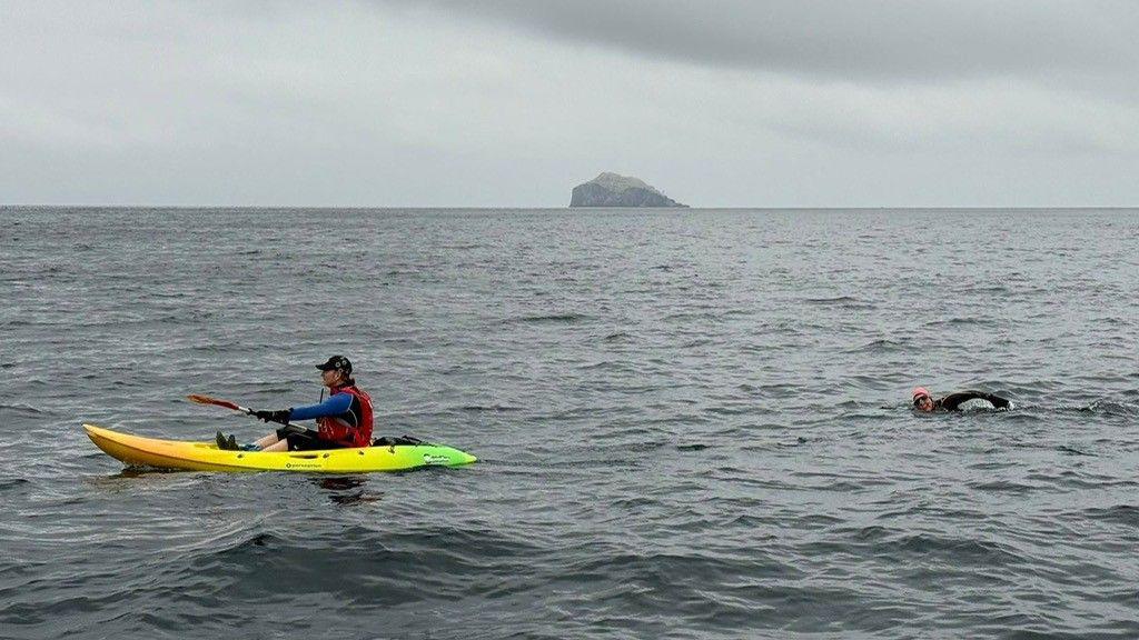 A man is paddling on a canoe, followed by a woman in a full wet suit and cap, swimming behind him. They are in a vast expanse of water, with only Bass Rock visible on the horizon. 