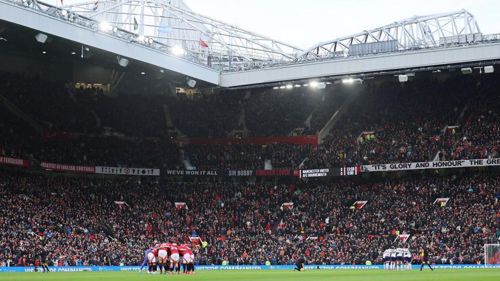 Manchester United players in a huddle in the middle of the pitch with a full Old Trafford in the background