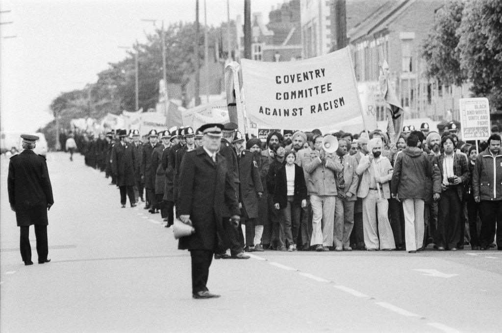 A long line of protesters hold a banner with the words Coventry Committee Against Racism whilst they are flanked by many police officers in uniform.