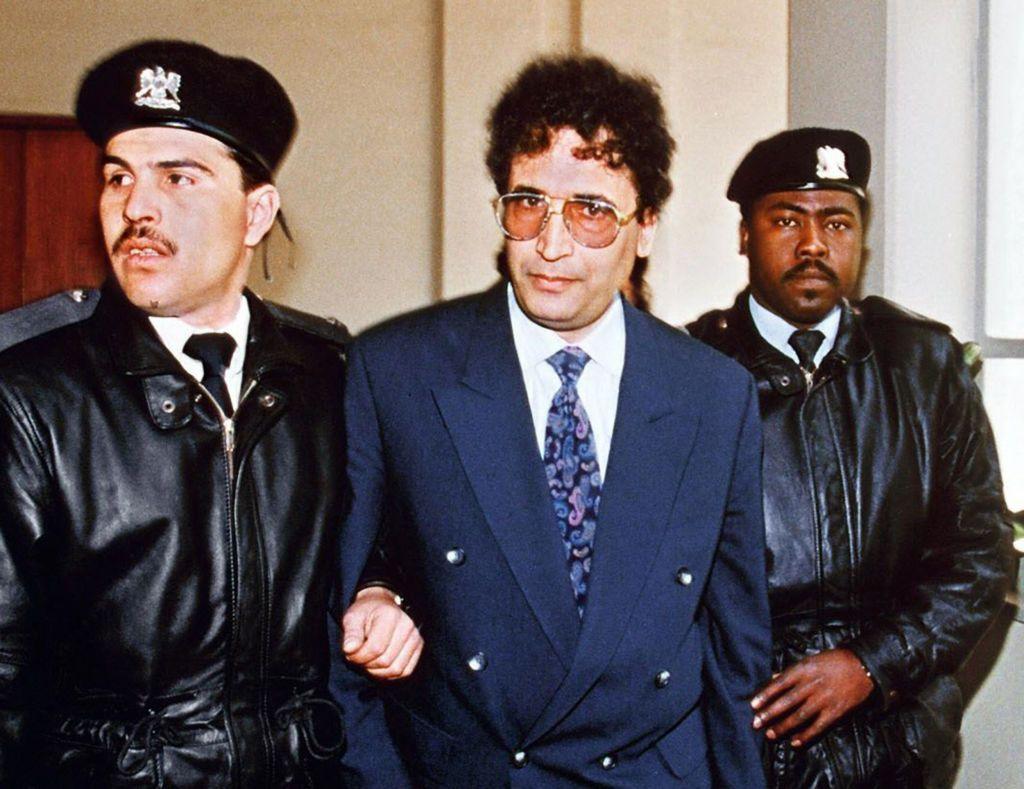 Abdelbasset al-Megrahi is escorted by two security officers in Tripoli in 1992. He is wearing a blue suit jacket, a white shirt, a patterned blue and purple tie and has large glasses. The officers are dressed in black coats and black berets.