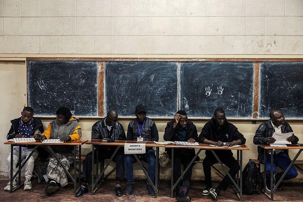 A group of officials sit behind trestle tables. Behind them are four school blackboards fixed to the wall.