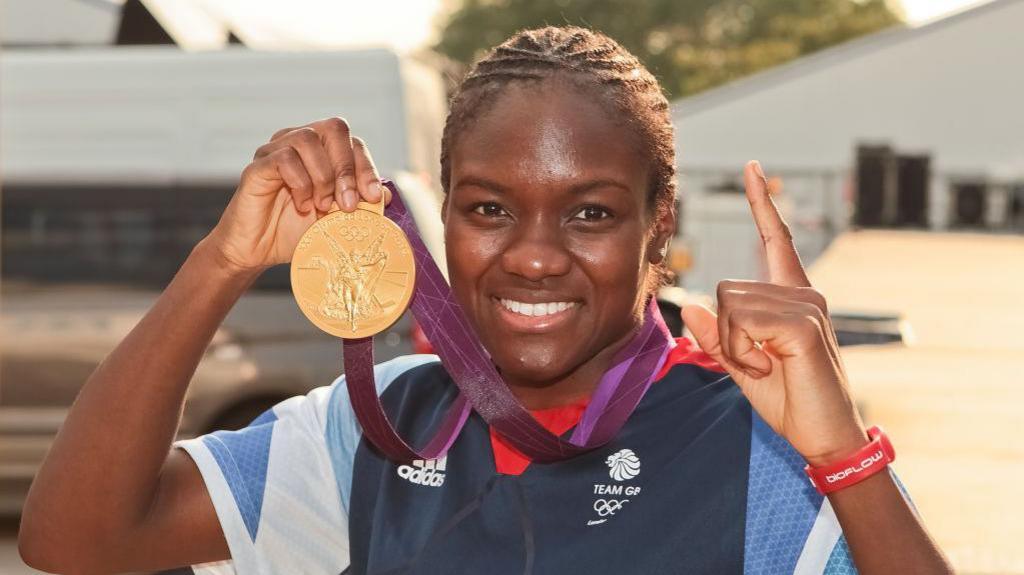 Nicola Adams holds the Olympic gold medal in one hand and her finger up in the other