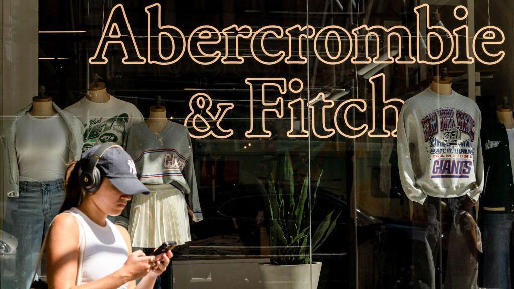 A shop window display of Abercrombie & Fitch featuring mannequins dressed in casual clothing. A woman is standing outside the store, looking at her phone.