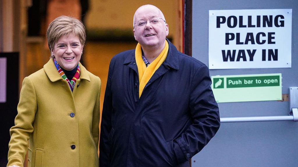 Nicola Sturgeon in a buttoned yellow coat is smiling outside a polling office. Peter Murrell is to her right, wearing a dark blue buttoned coat and a bright yellow scarf. They are smiling at the camera.