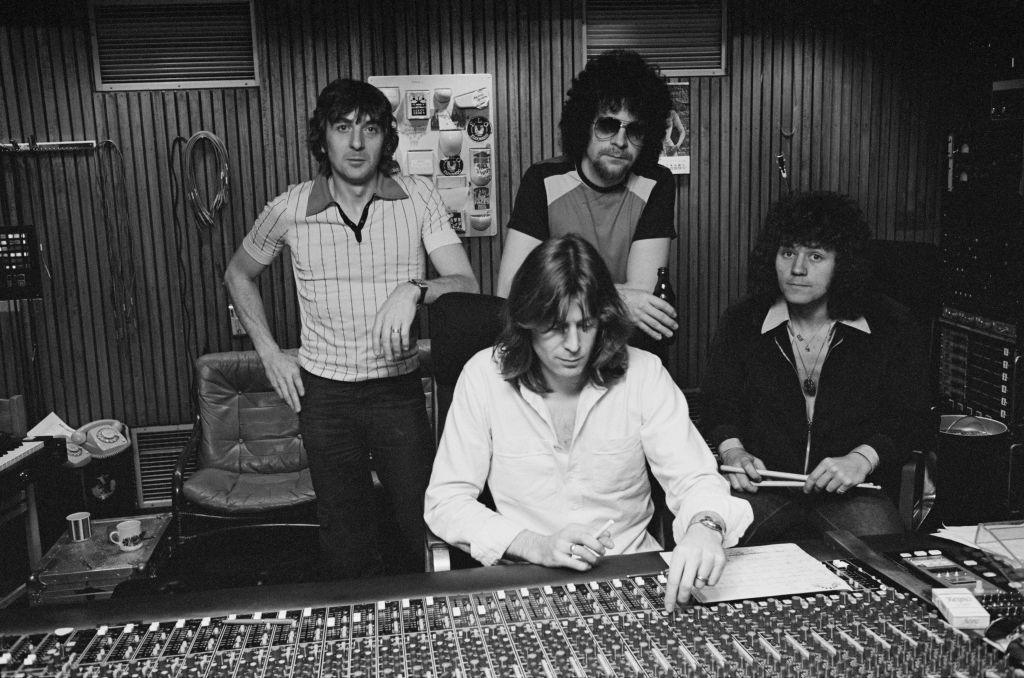 A black and white photo of four men in a recording studio. The men are wearing 1970s-style clothes and have long hair. The man at the front of the group is sitting down and sliding a button on the recording desk. One of the other men is holding drumsticks.