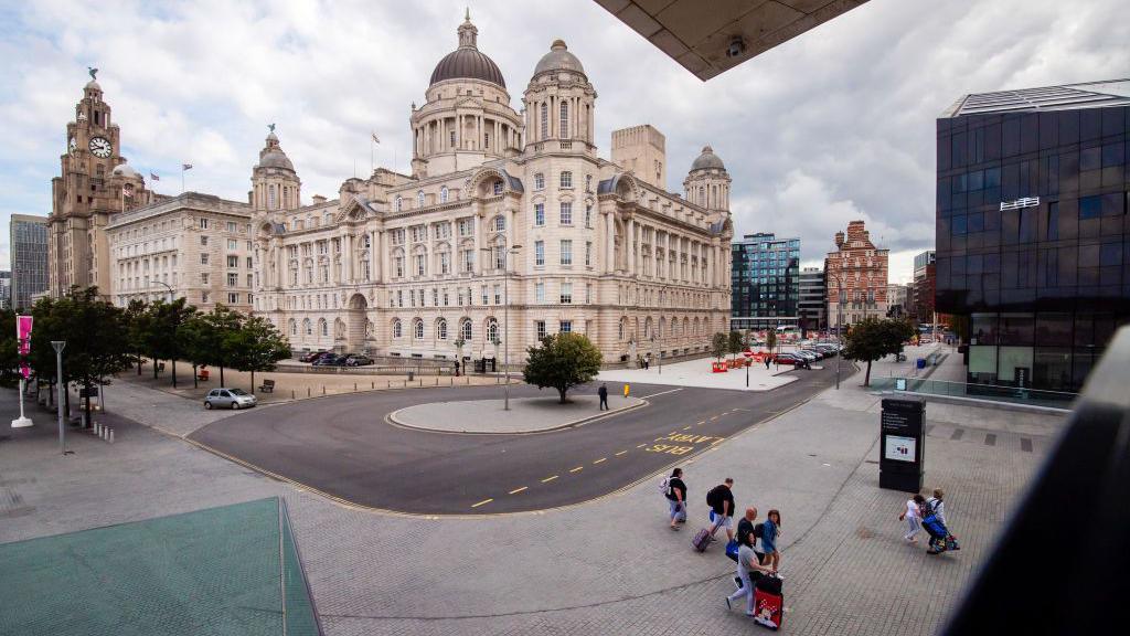 Visitors carry baggage on the Pier Head, with the Royal Liver Building, the Cunard Building and The Port of Liverpool building, known collectively as the Three Graces