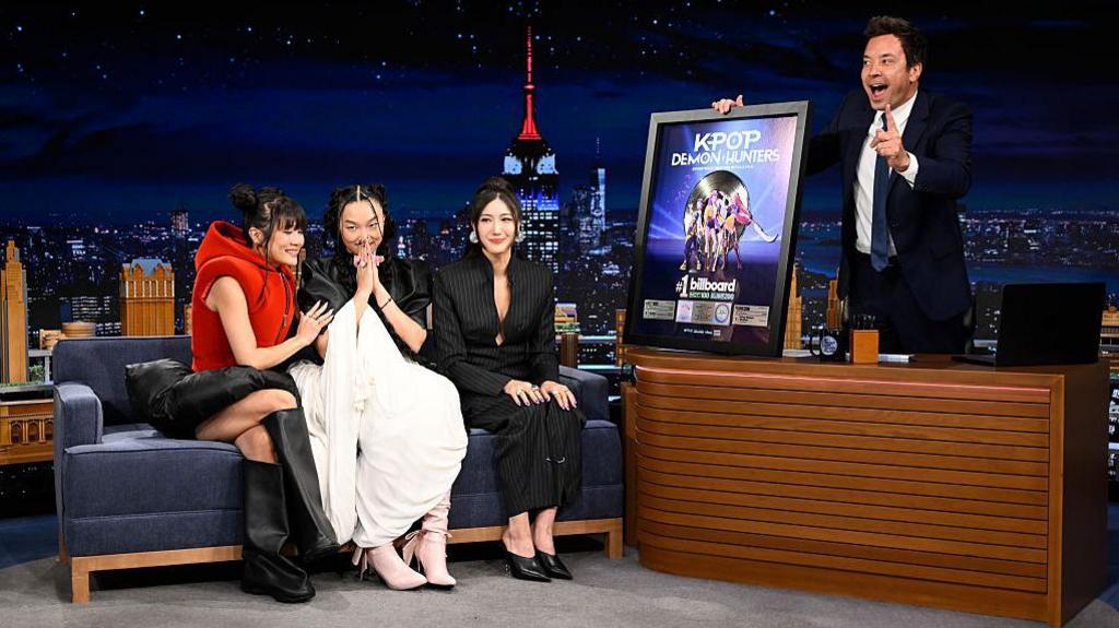 Jimmy Fallon presenting a platinum record for the kpop demon hunters soundtrack to EJAE, Audrey Nuna and Rei Ami sitting on the show's sofa