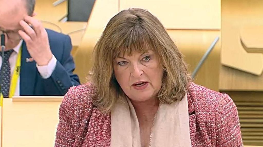 Fiona Hyslop, wearing a pink jacket and pink scarf, speaks in the Holyrood chamber