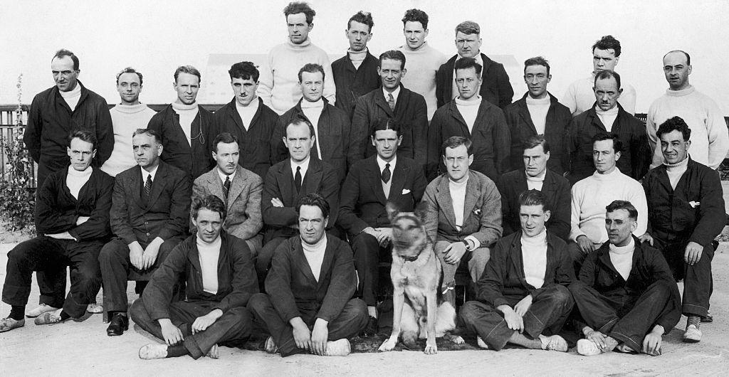 Officers and members of the crew of the R101 pose for photographs after another day of flight trials. There about 30 men in four rows. The men in the front row are sitting on the ground. Some are wearing suits and ties, others are in boiler suits and jumpers. There is a large dog, which looks like an alsatian, in the middle of the front row. 