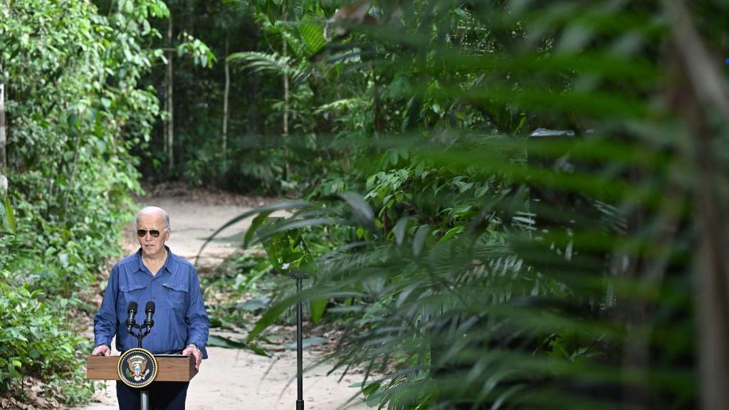 Joe Biden makes statements to the news media from a podium in the Amazon rainforest. He is surrounded by greenery and tropical trees and brush.  