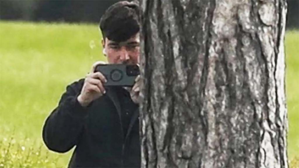 A man in a dark jacket and blue jeans stands partially concealed by a tree trunk and holds a phone up in two hands as if filming and has a white earphone in his right ear