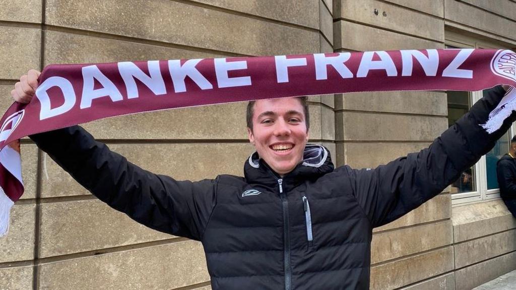 Josef is wearing a black puffer jacket and is holding up a scarf that says 'Danke Franz'