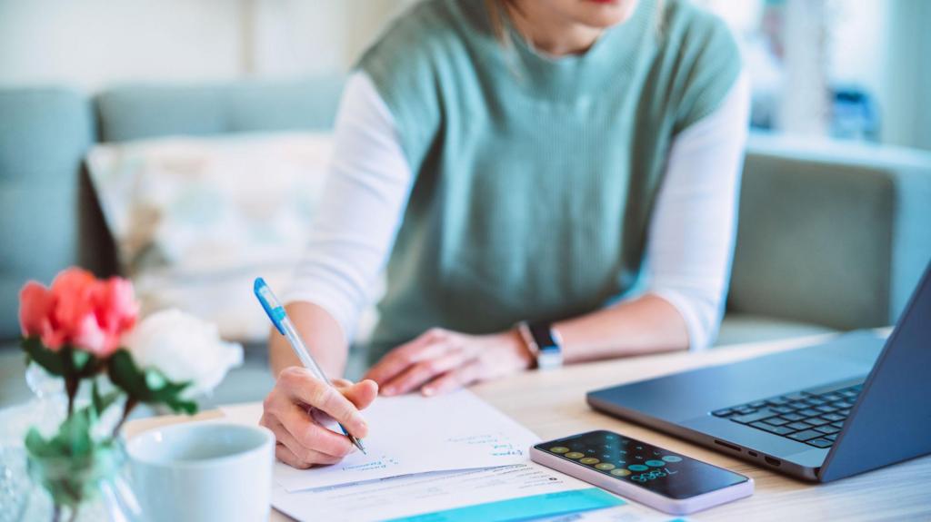 A woman in a blue top sits at a wooden table looking at something on her laptop, a phone next to it with the calculator app open. The woman is writing down something on a piece of paper