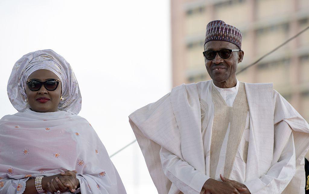 Buhari stands next his wife. They both wear dark sunglasses and white outfits.