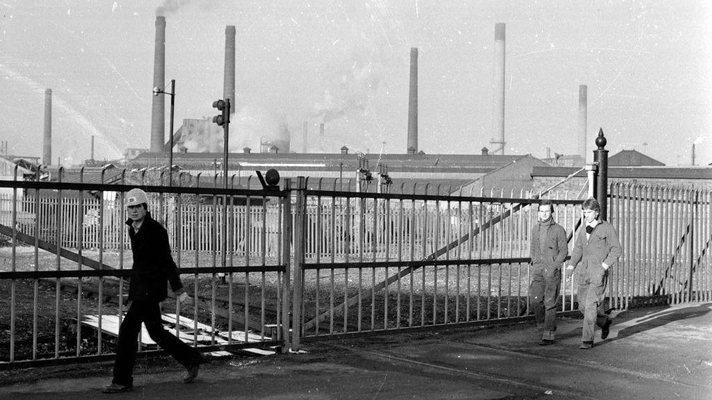 A black and white photo of Corby's steelworks. It is a huge factory with several chimneys. It is in the distance behind a fence. A man in a hard hat looks at the camera as he walks in front of the fence. Two other men in boiler suits are walking past a short distance behind him.