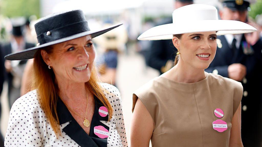 Sarah Ferguson and Princess Eugenie at Royal Ascot 2025. Ferguson wears a white dress with black spots and a black hat. Eugenie, on her left, wears a brown sleeveless dress and a white hat. Both are wearing two pink badges. 