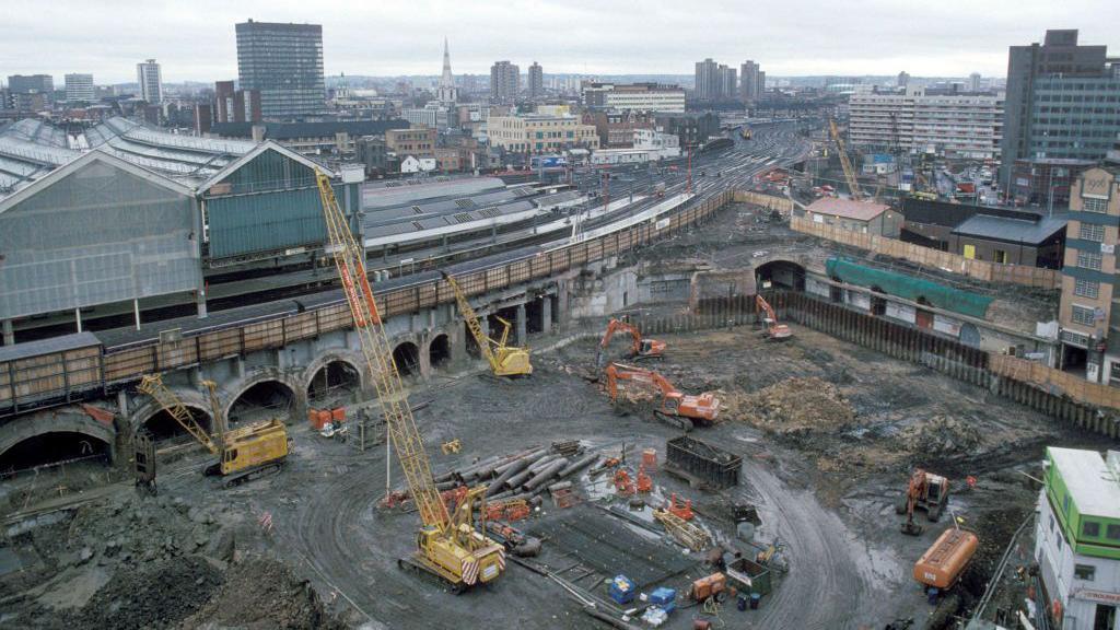 A building site behind Waterloo station with cranes, diggers and other construction vehicles seen. The train tracks are seen above the site and going off into the distance, with the city visible beyond it