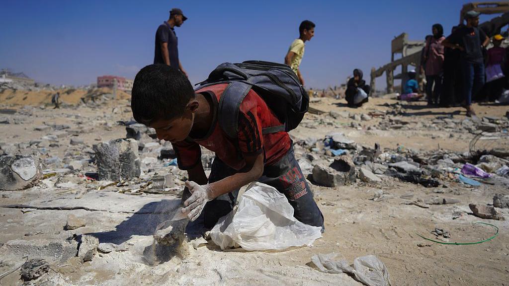 A child is on his knees trying to collect flour from the ground in Gaza. The floor is littered with rubbish and rocks and other people walk by him in the background.