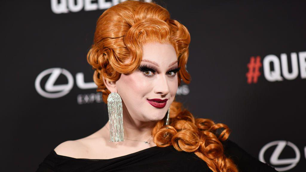 Drag Queen Jinkx Monsoon - in glamorous Hollywood make-up and red lips, with a bright ginger curled wig and huge diamante earrings. She is smiling at the camera while attending the Queerties event in 2024.