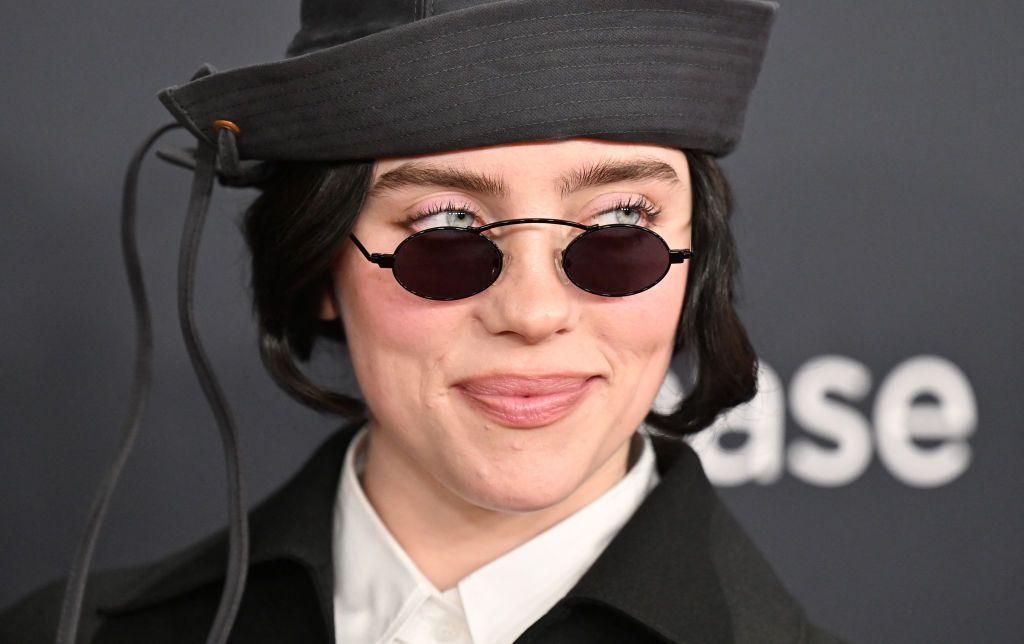 Billie Eilish a young woman with dark shiny hair wears a black and white outfit including a black sailor-type hat, with black circle frame sunglasses she peeks over to the side with a small smile.