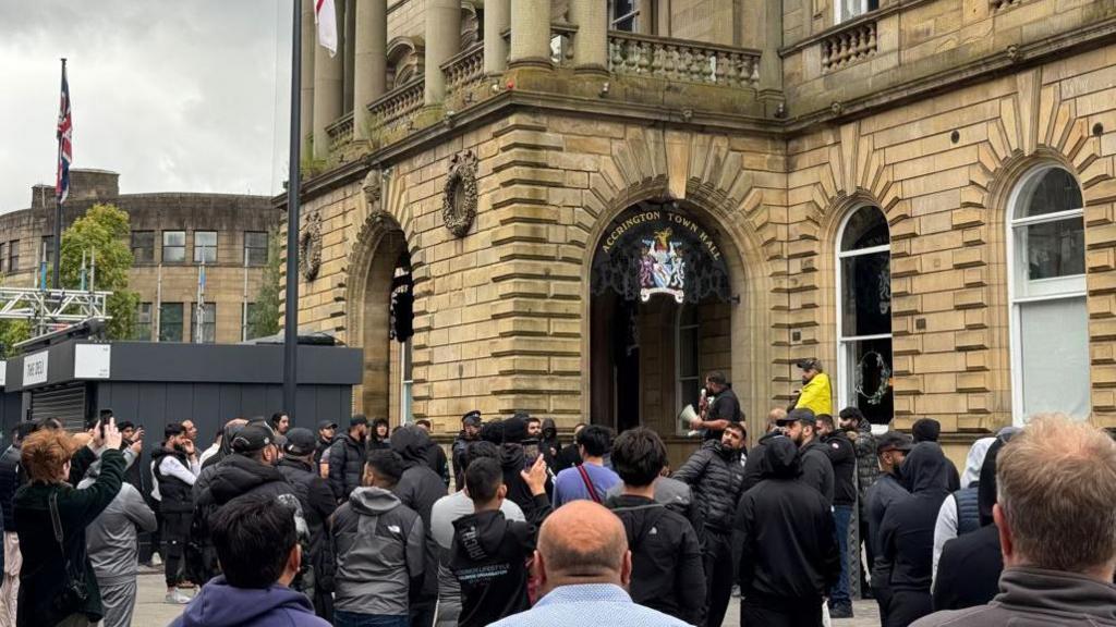 Crowds gather in Accrington outside the Town Hall - a man is on a megaphone and some people hold up mobile phones filming the event