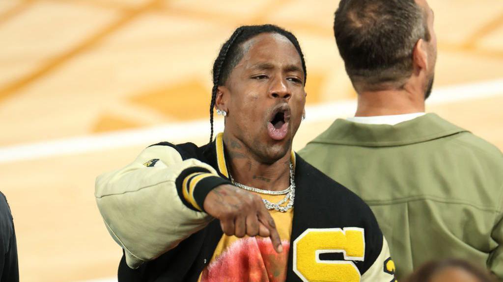 Travis Scott wears a black and yellow varsity jacket with a large yellow "S" on the left breast. He's gesturing and shouting, pointing downwards, in an apparent celebration.